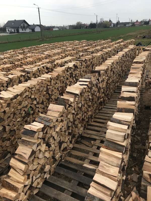 Rzędami ułożone drewno kominkowe na paletach, widok z perspektywy, w tle budynek mieszkalny i pole.