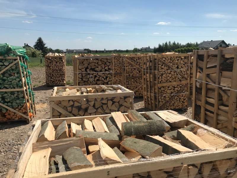 Kilkanaście drewnianych skrzyń wypełnionych pociętym drewnem opałowym, niektóre z korą, ustawionych na zewnątrz w słoneczny dzień.