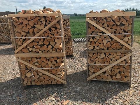 Dwa kontenery z pociętym drewnem ułożone na żwirowym podłożu; w tle zielona trawa i błękitne niebo.