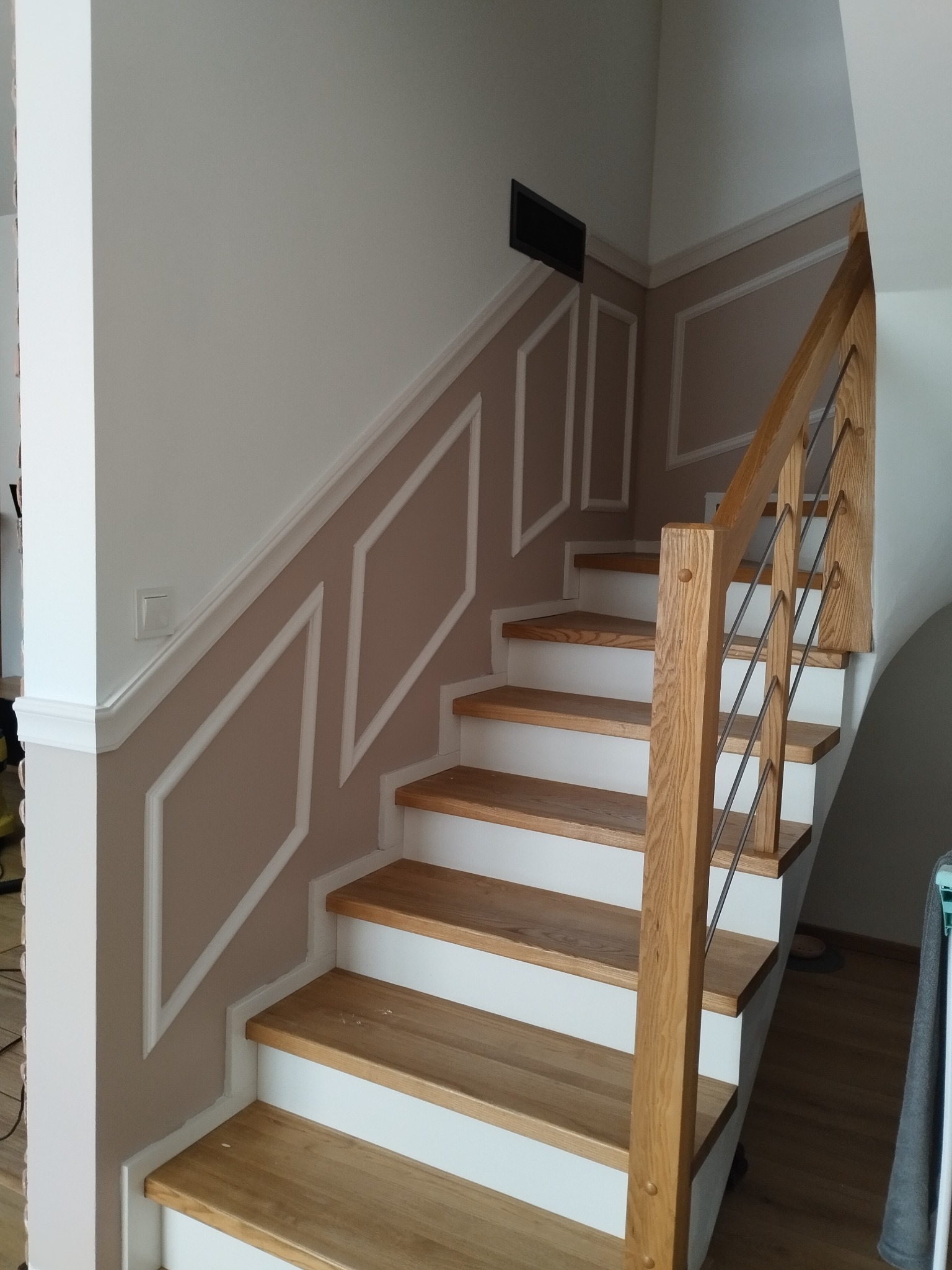 Drewniane schody z białymi podstopnicami i metalową balustradą. Ściany zdobione sztukaterią w geometryczne wzory. Widok z dołu na jasne, eleganckie wnętrze.