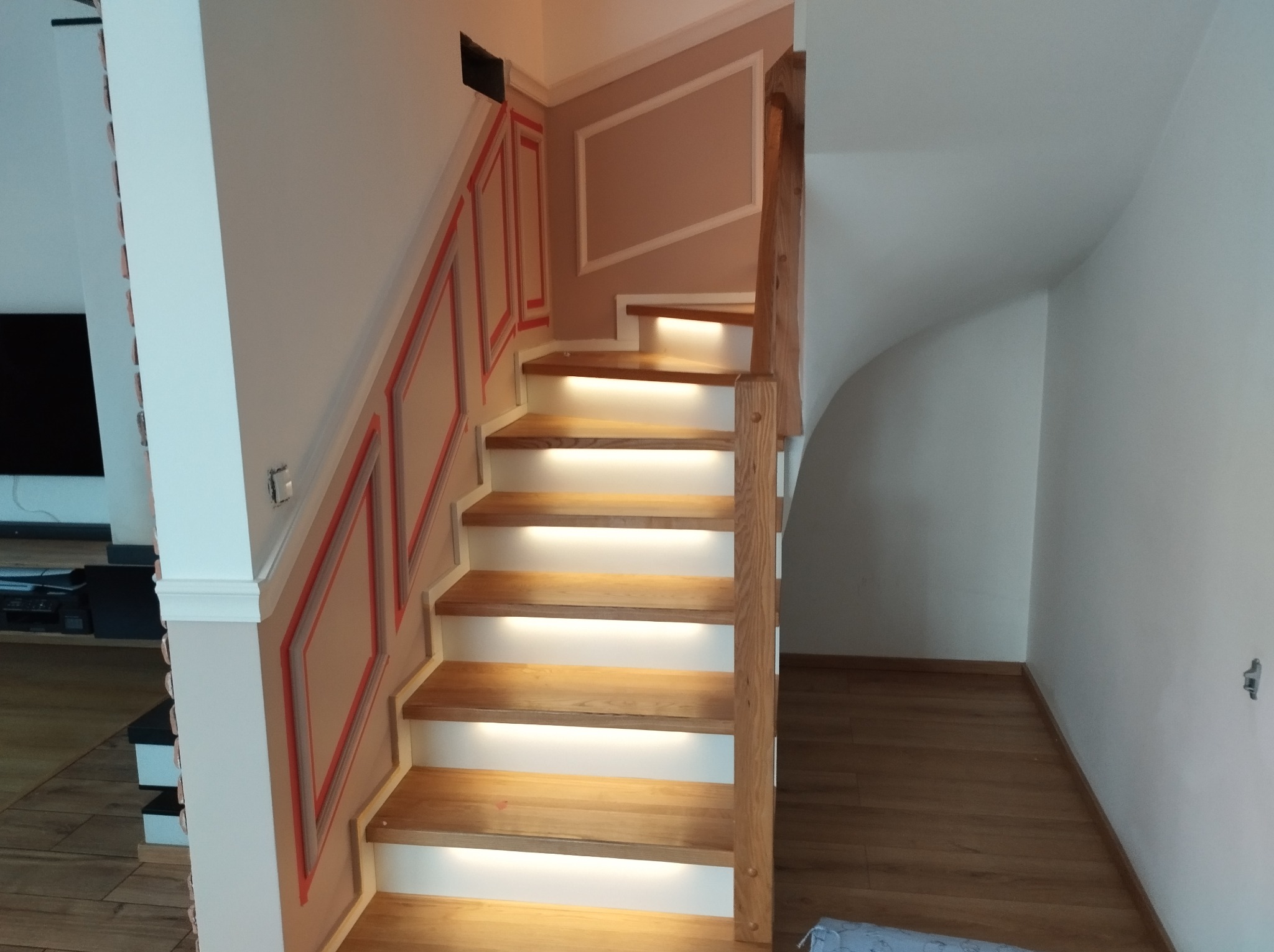 Drewniane schody z podświetleniem LED, ozdobne panele ścienne w kolorze beżowym z czerwonymi obramowaniami. Widok z dołu, jasne oświetlenie.