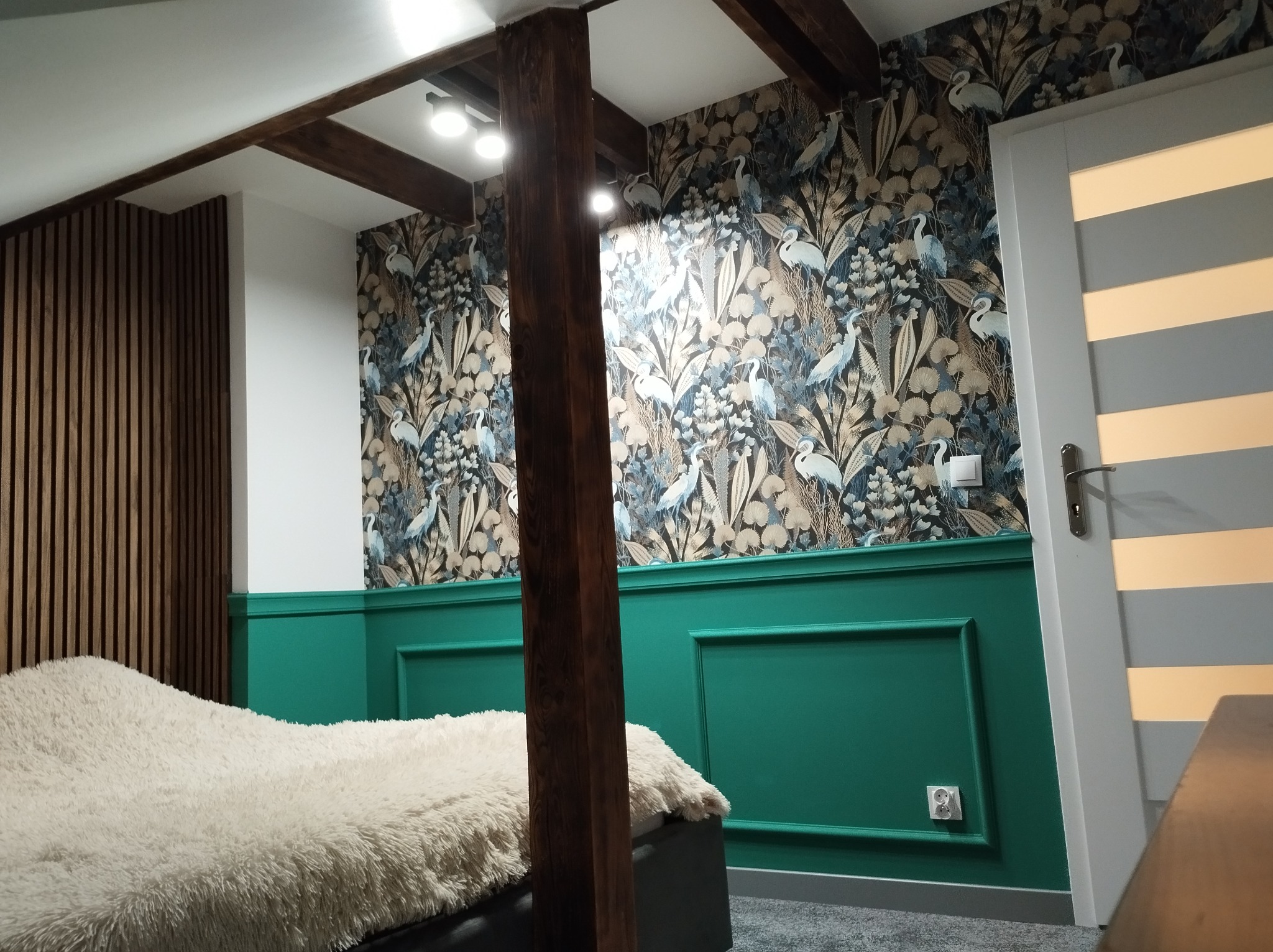 Stylowa sypialnia z tapetą w motywy roślinne i ptasie, zieloną boazerią, drewnianymi belkami i puszystym kocem na łóżku. Widoczny fragment drzwi z poziomymi paskami.