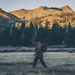 Luke Bochenek - Mężczyzna w outdoorowej odzieży i z plecakiem idzie przez trawiaste pole w kierunku lasu i gór w oddali, wczesnym rankiem.