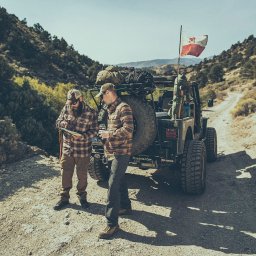 Luke Bochenek - Dwóch mężczyzn w plenerze w stylu lifestyle/fashion, obok terenowego jeepa z bagażami i flagą, analizujących mapę na tle górskiego krajobrazu.