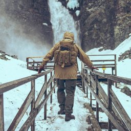 Luke Bochenek - Osoba w brązowej kurtce i z plecakiem idzie po drewnianym mostku w kierunku wodospadu w zimowym krajobrazie.