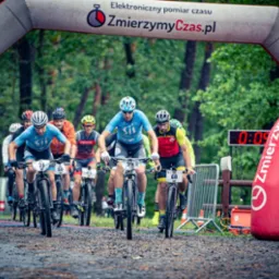 Grupa kolarzy górskich rywalizuje w reportażu z wyścigu, przejeżdżając pod łukiem startowym z elektronicznym pomiarem czasu na leśnym terenie.