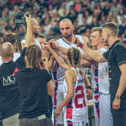 Luke Bochenek - Drużyna koszykarska w białych strojach z czerwonymi akcentami, złączona dłońmi w kręgu, otoczona przez fotografa z aparatem i trenera, na tle rozmazanego tłumu kibiców.