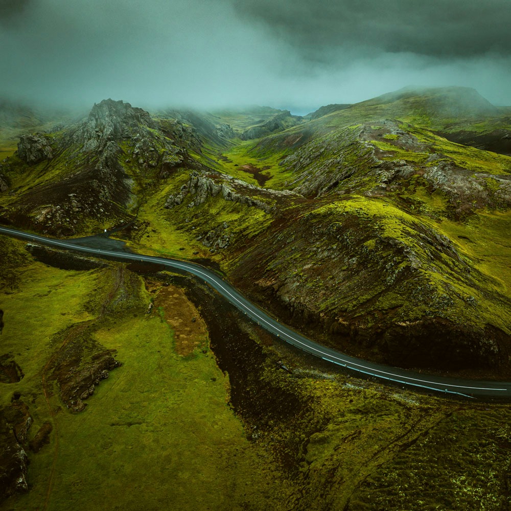 Pejzaż z lotu ptaka: Kręta droga przecinająca zielone, mchem porośnięte wzgórza i skaliste zbocza w mglistej dolinie.