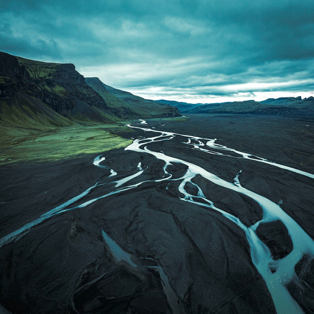 Widok z drona na islandzki krajobraz z rzeką wijącą się przez czarną, wulkaniczną ziemię, z zielonymi wzgórzami w tle i pochmurnym niebem.