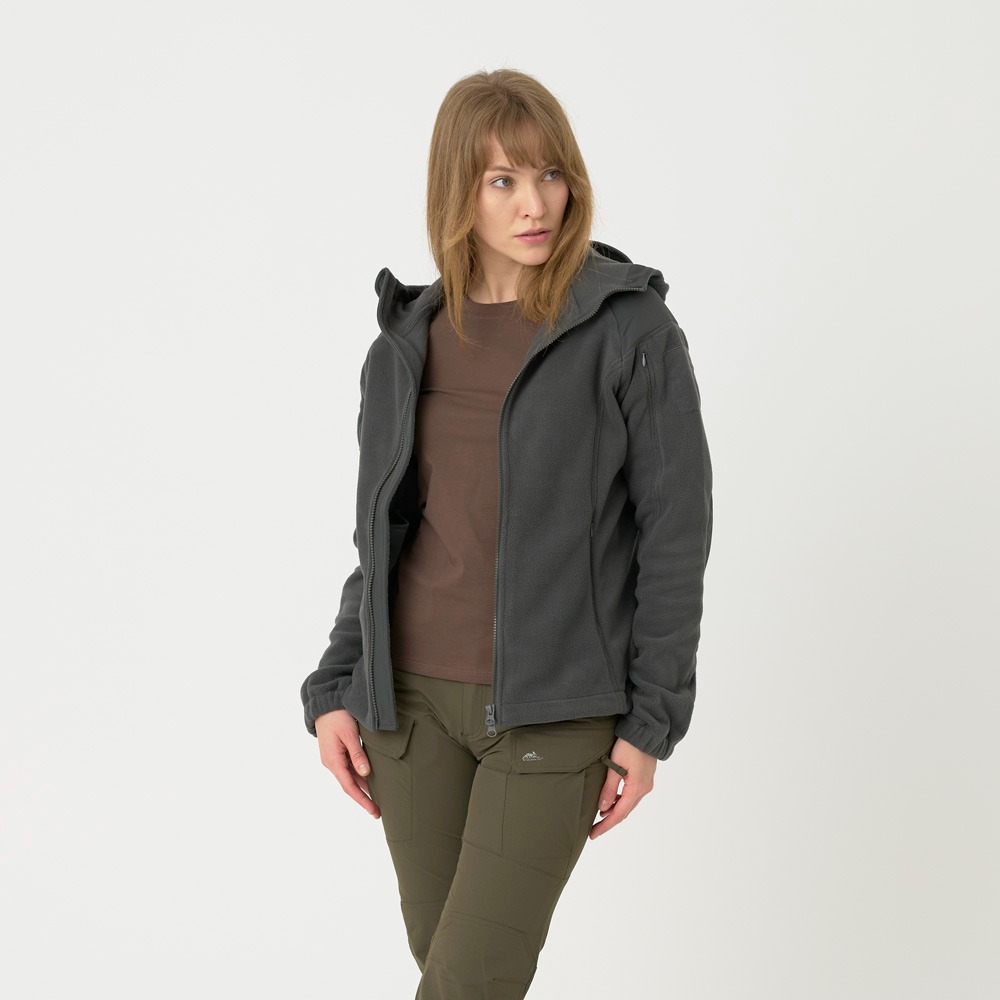 Kobieta prezentuje odzież outdoorową: szarą bluzę polarową z kapturem i oliwkowe spodnie cargo na białym tle, wzrok skierowany w lewo.