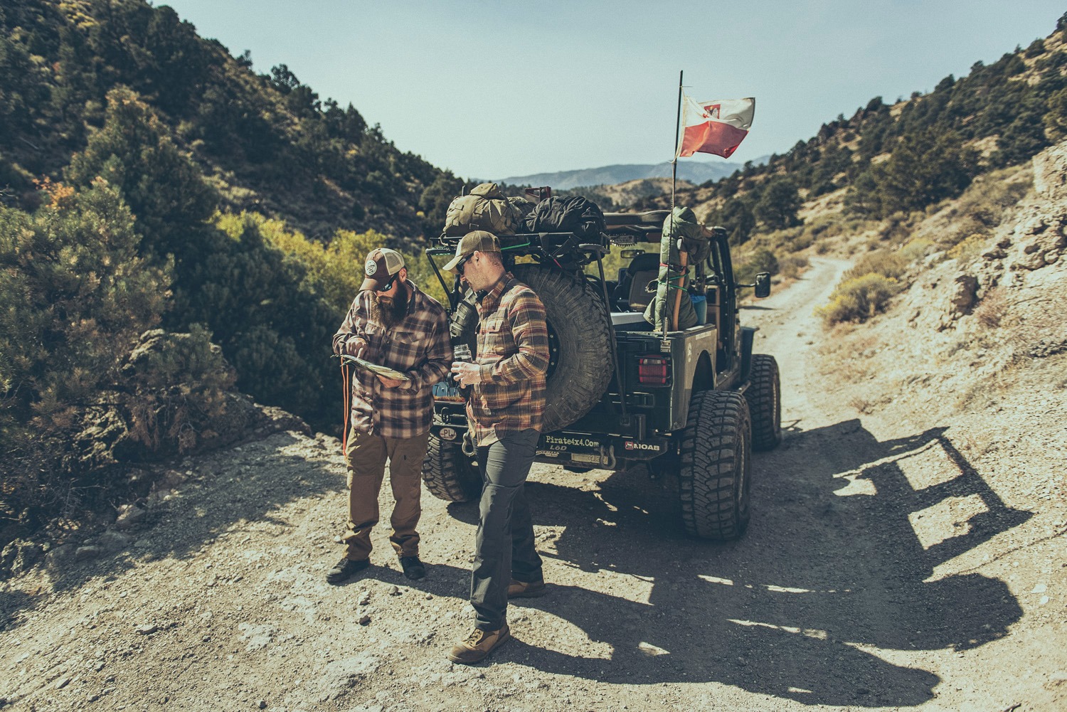Dwóch mężczyzn w plenerze w stylu lifestyle/fashion, obok terenowego jeepa z bagażami i flagą, analizujących mapę na tle górskiego krajobrazu.
