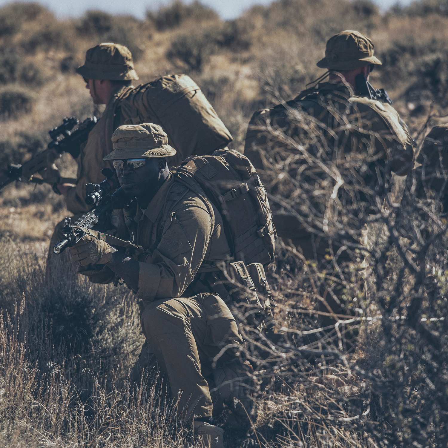 Grupa żołnierzy w kamuflażu, z karabinami i plecakami, w pozycji gotowości w suchym, trawiastym terenie.
