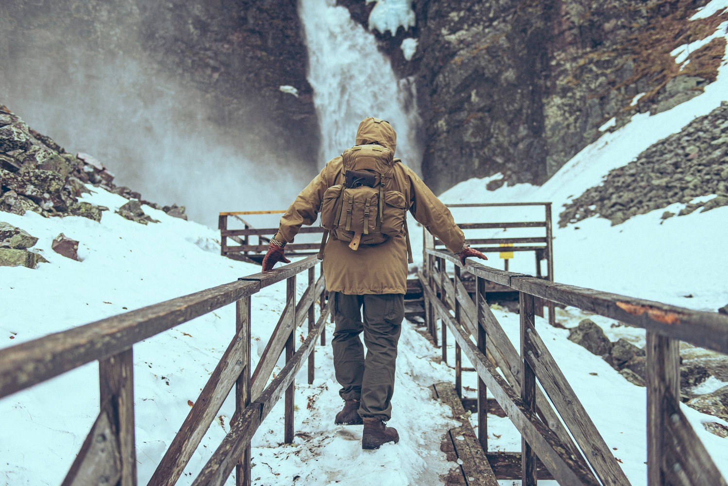 Osoba w brązowej kurtce i z plecakiem idzie po drewnianym mostku w kierunku wodospadu w zimowym krajobrazie.