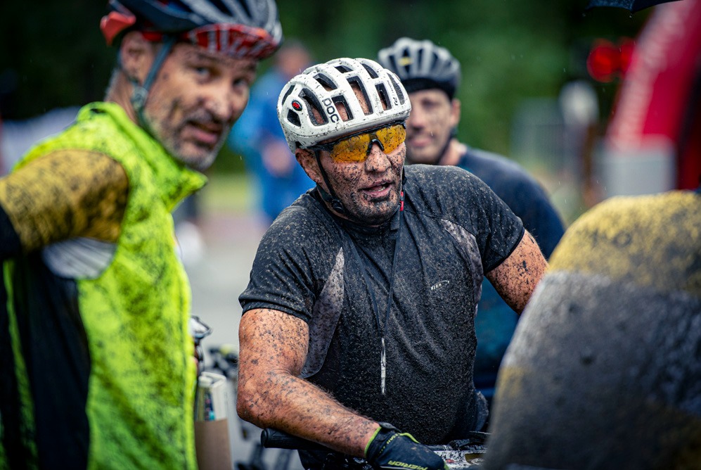 Umazany błotem rowerzysta w kasku i okularach, reportaż z zawodów MTB, w tle inni zawodnicy i rowery.