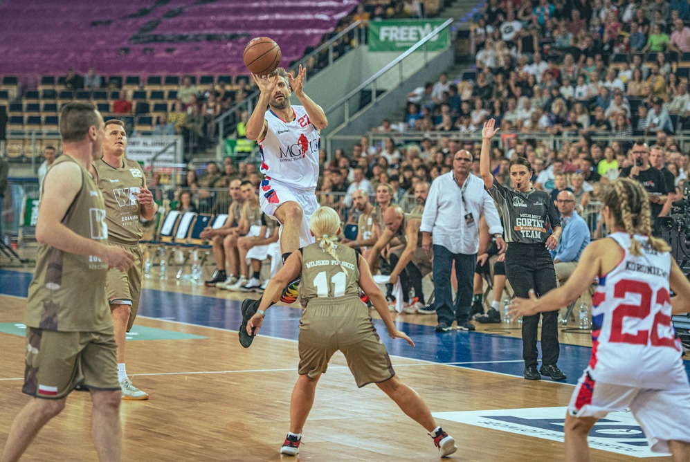 Mężczyzna w białej koszulce z numerem 27 skacze, aby rzucić piłkę do kosza podczas meczu koszykówki; w tle widoczna publiczność i sędzia.