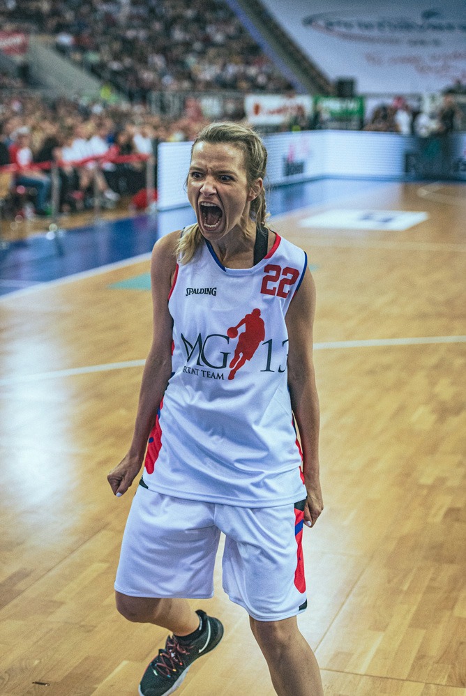 Rozemocjonowana koszykarka w białym stroju z numerem 22 i logo drużyny MG 13, krzycząca na parkiecie podczas meczu, widownia w tle.