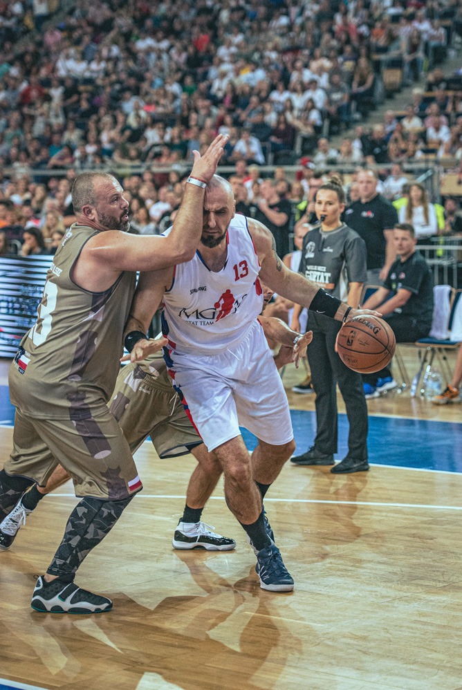 Mecz koszykówki: zawodnik w białym stroju z numerem 13 blokowany przez przeciwnika w brązowym stroju na tle trybun wypełnionych kibicami.