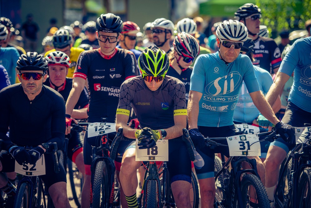 Grupa kolarzy górskich z numerami startowymi na rowerach, czekających na rozpoczęcie wyścigu, w pełnym słońcu.