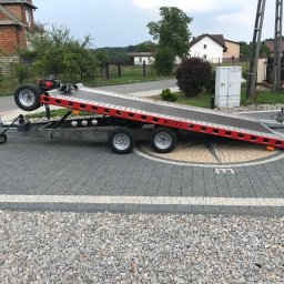TRADE Sikora Andrzej - Czerwono-szara przyczepa z najazdem, zaparkowana na kostce brukowej z widocznym napisem 'WIOŁA', w tle domy i słupy elektryczne.
