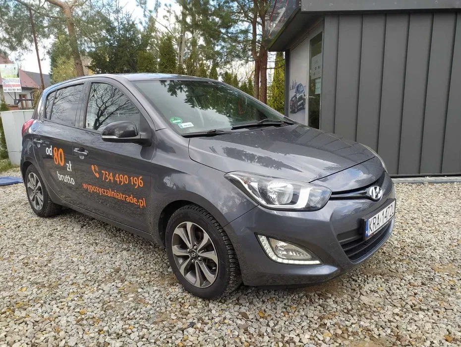 Szary Hyundai i20 z naklejką reklamową 'od 80 zł brutto 734 490 196 wypozyczalniatrade.pl' zaparkowany na żwirowym parkingu przed nowoczesnym budynkiem.