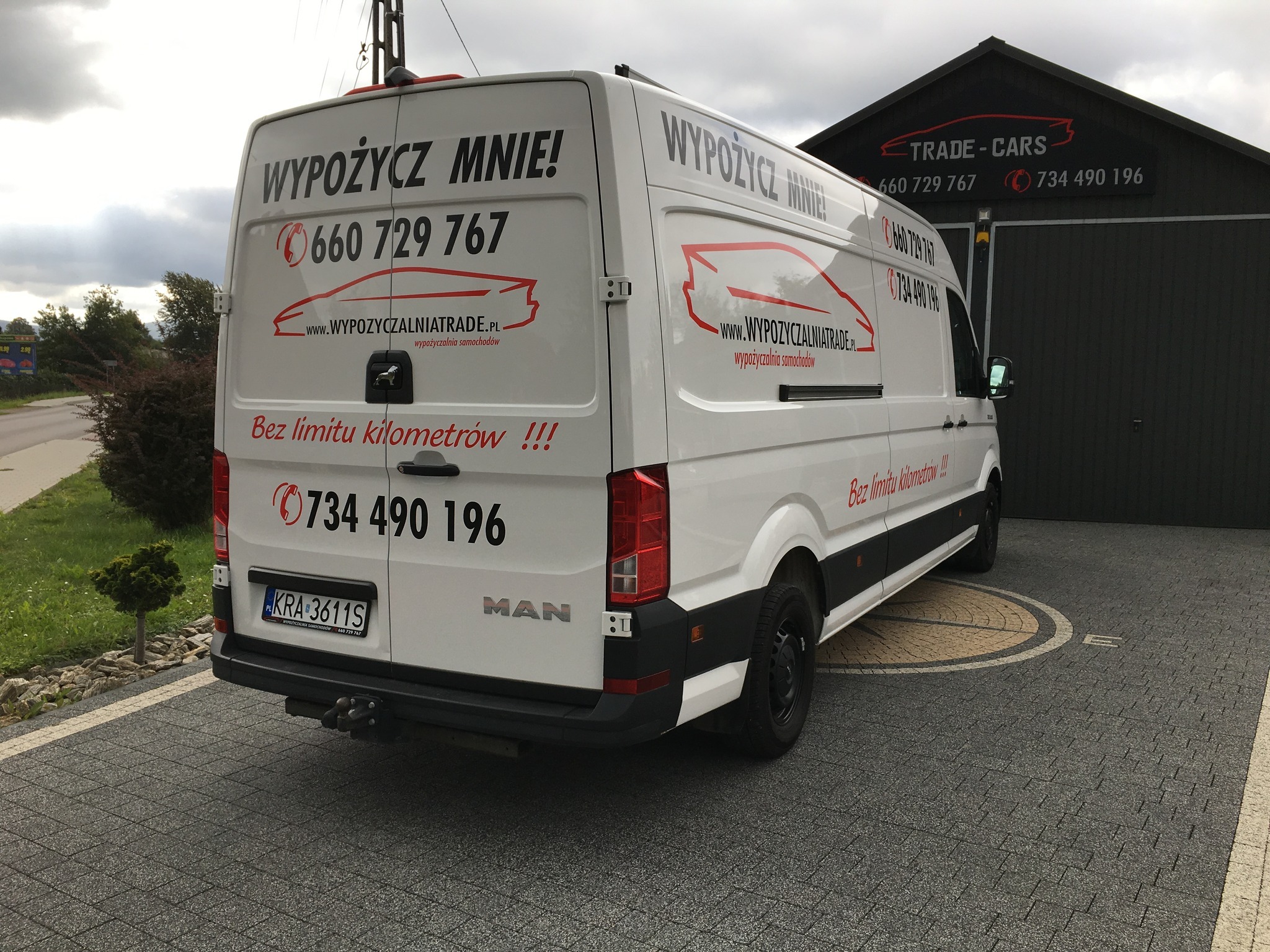Biały samochód dostawczy marki MAN z napisem 'WYPOŻYCZ MNIE!' i numerami telefonów, zaparkowany na kostce brukowej przed budynkiem firmy 'TRADE-CARS', oferujący wynajem samochodów bez limitu...
