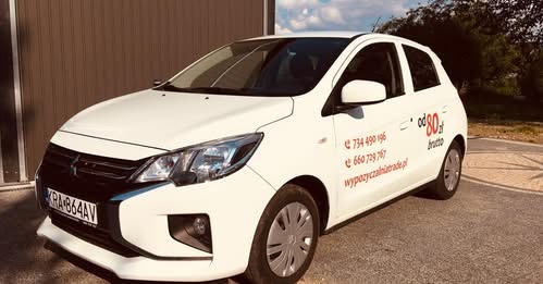 Biały samochód osobowy z naklejkami informacyjnymi o cenie wynajmu i numerach kontaktowych, zaparkowany na zewnątrz w słoneczny dzień.