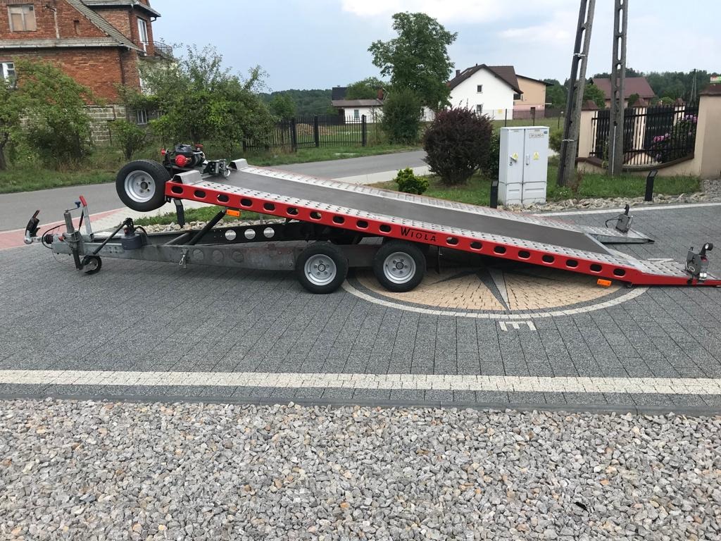 Czerwono-szara przyczepa z najazdem, zaparkowana na kostce brukowej z widocznym napisem 'WIOŁA', w tle domy i słupy elektryczne.