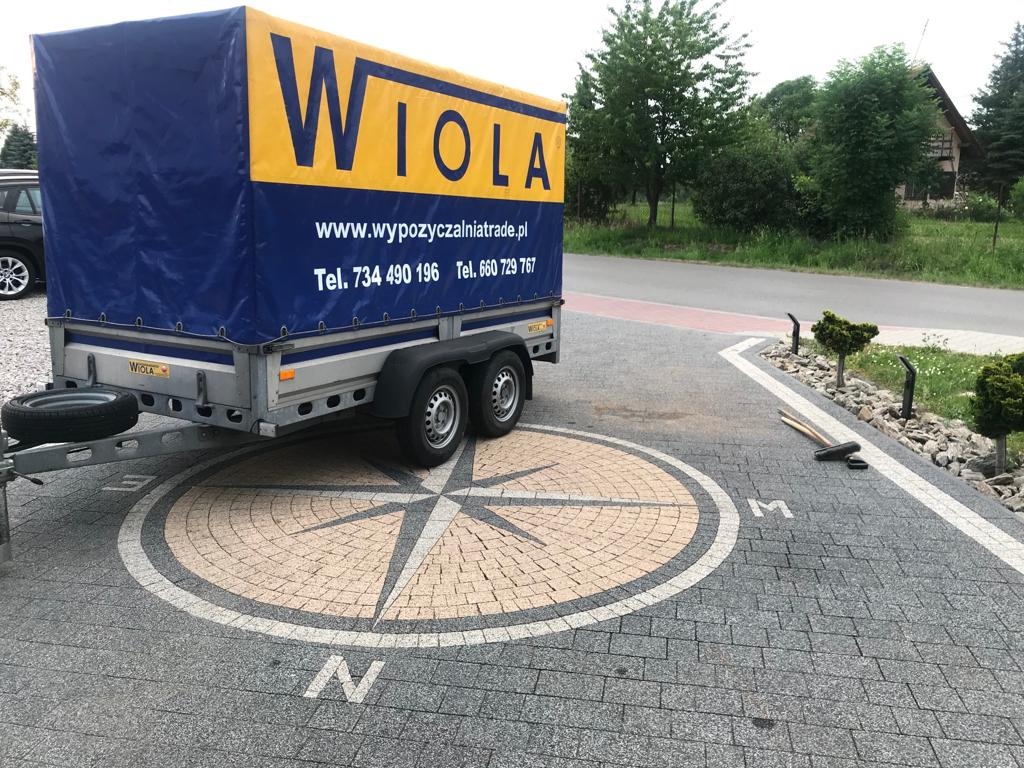 Przyczepa z plandeką z logo firmy 'Wiola' na tle kostki brukowej z motywem kompasu, widok z boku, z zaparkowanym samochodem w tle.