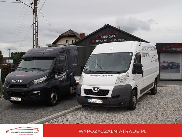 Czarny samochód typu laweta marki Iveco i biały bus marki Peugeot na żwirowym placu przed budynkiem z napisem TRADE-CARS i WYPOZYCZALNIATRADE.PL.