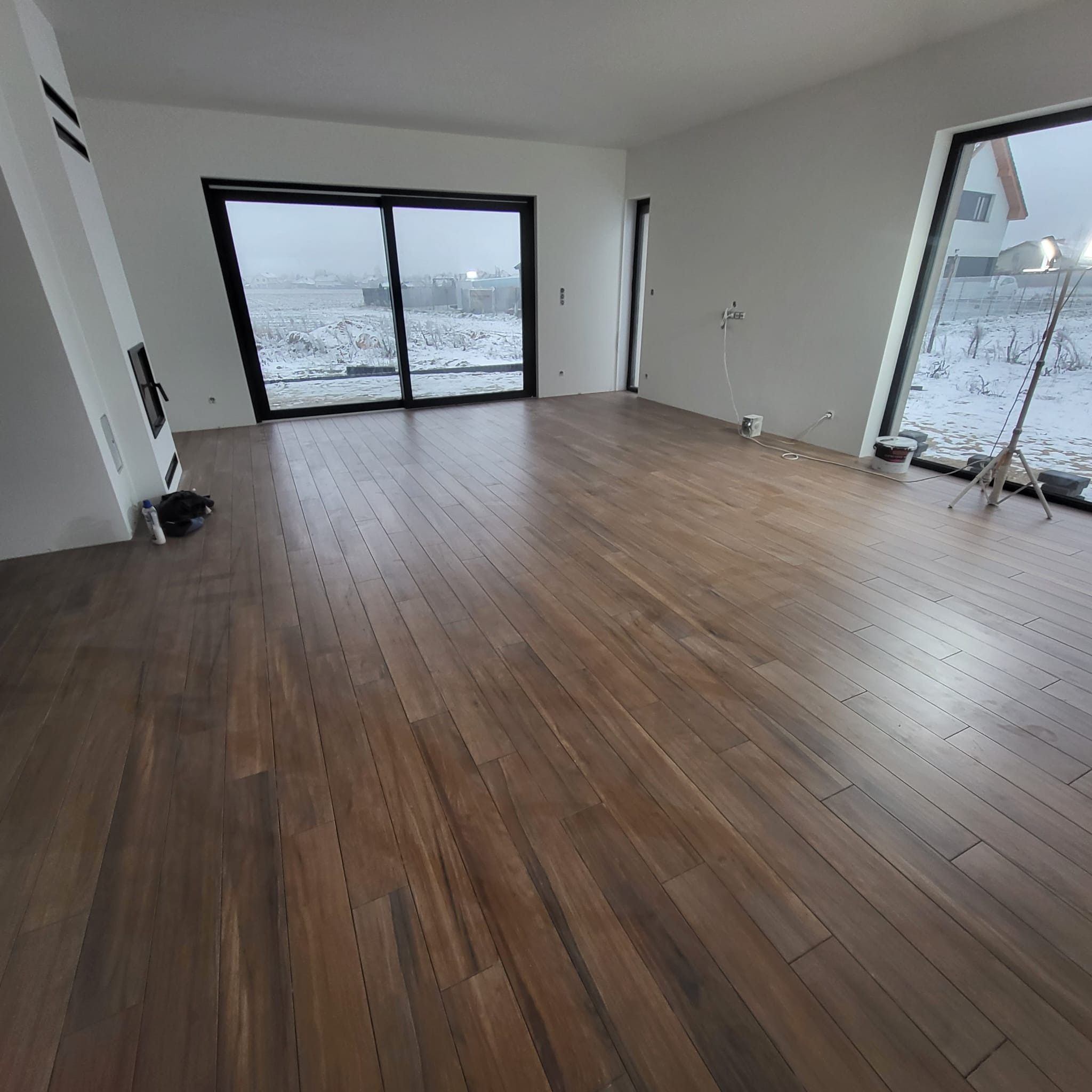 Salon w stanie deweloperskim z ułożoną podłogą z paneli drewnopodobnych, widoczne duże okna z widokiem na zimowy krajobraz i lampa budowlana.