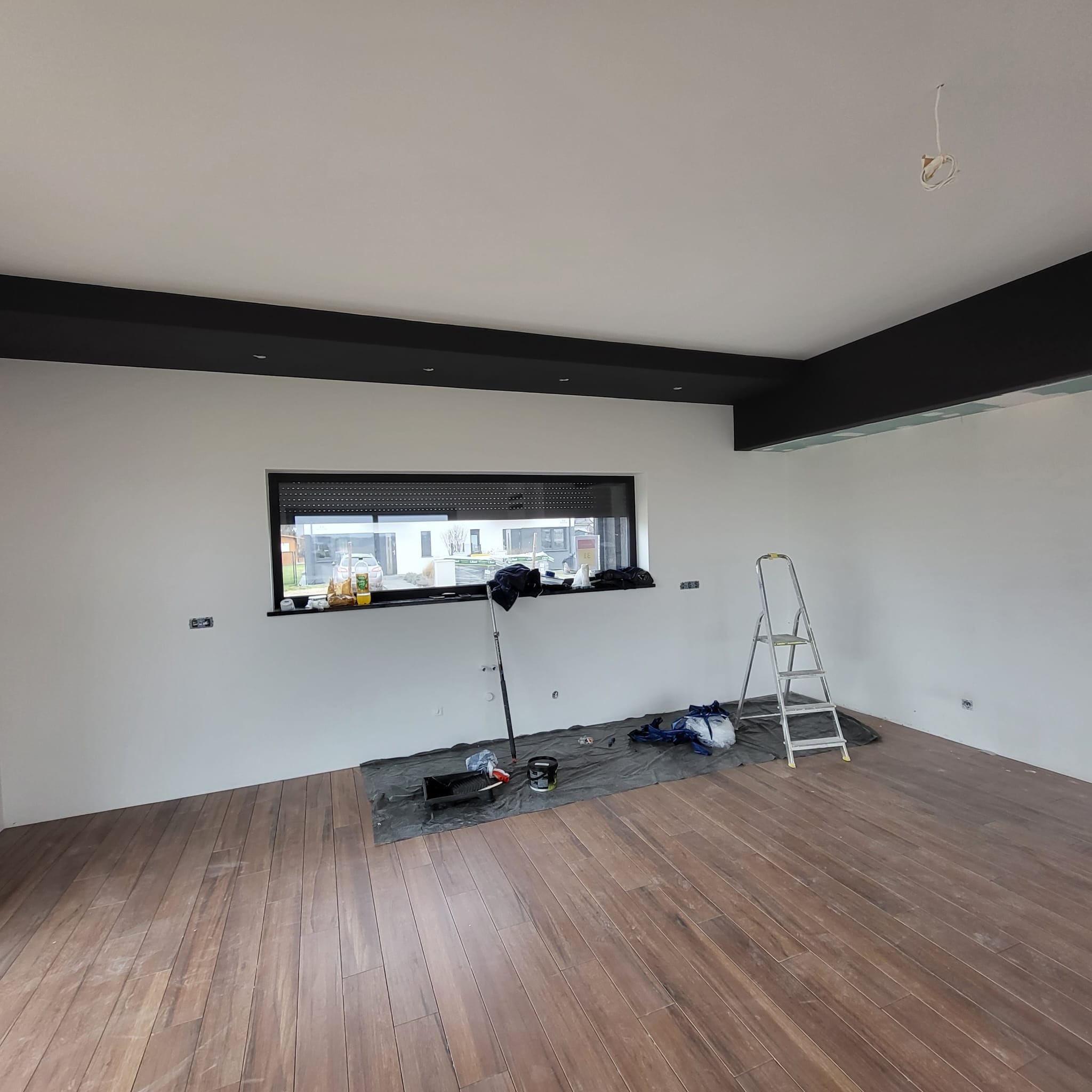 Pokój w trakcie malowania: białe ściany, czarne belki sufitowe, drewnopodobne panele podłogowe, drabina, akcesoria malarskie na folii ochronnej. Widoczne niedokończone podłączenie elektryczne...