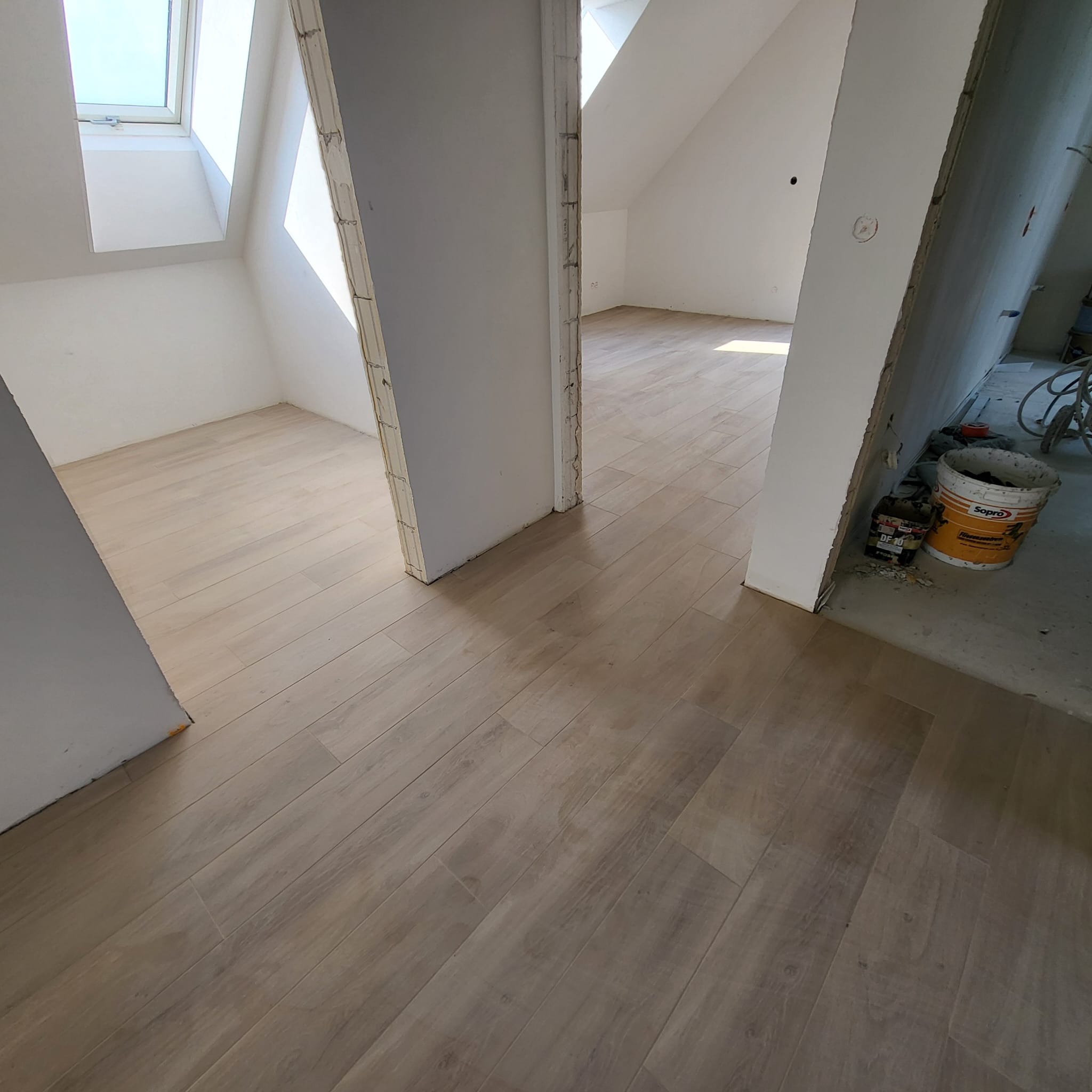 Ułożone panele podłogowe imitujące drewno w jasnym odcieniu, widok z perspektywy korytarza w kierunku poddasza, fragment puszki po kleju do paneli Sopro DF 10 przy ścianie.