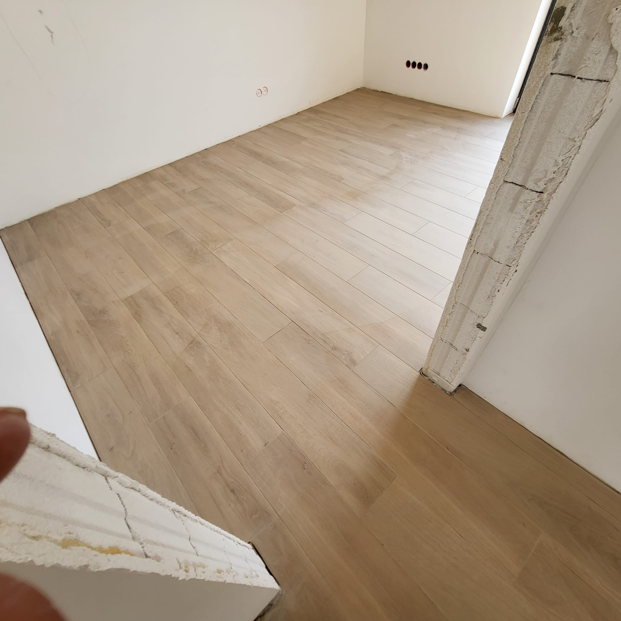 Ułożone panele podłogowe w jasnym odcieniu drewna, widoczne niedokończone obramowanie drzwi z surowego betonu i białe ściany z gniazdkami elektrycznymi.