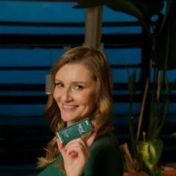 Uśmiechnięta blondynka prezentuje zielone opakowanie suplementu diety Terra+ Minerals na tle roślin i niebieskich paneli.