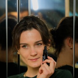 Terra Plus - sklep z suplementami diety - kwas fulwowy - Kobieta z zielonymi oczami i kolczykiem w nosie trzymająca buteleczkę olejku Terra Alkaline na tle lustra.