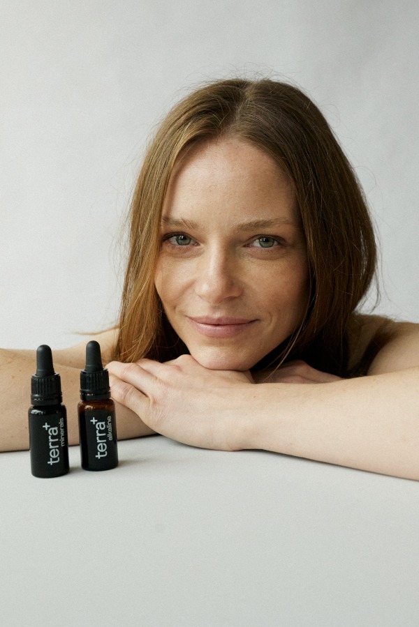 Kobieta z jasnymi włosami opiera podbródek na dłoniach, obok niej dwie buteleczki z kroplomierzem marki Terra+ minerals i Terra+ alkaline.