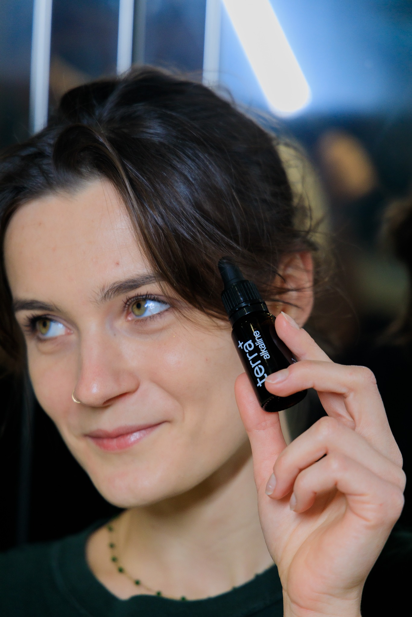 Uśmiechnięta kobieta z kolczykiem w nosie prezentuje czarną buteleczkę z kroplomierzem Terra Alkaline.