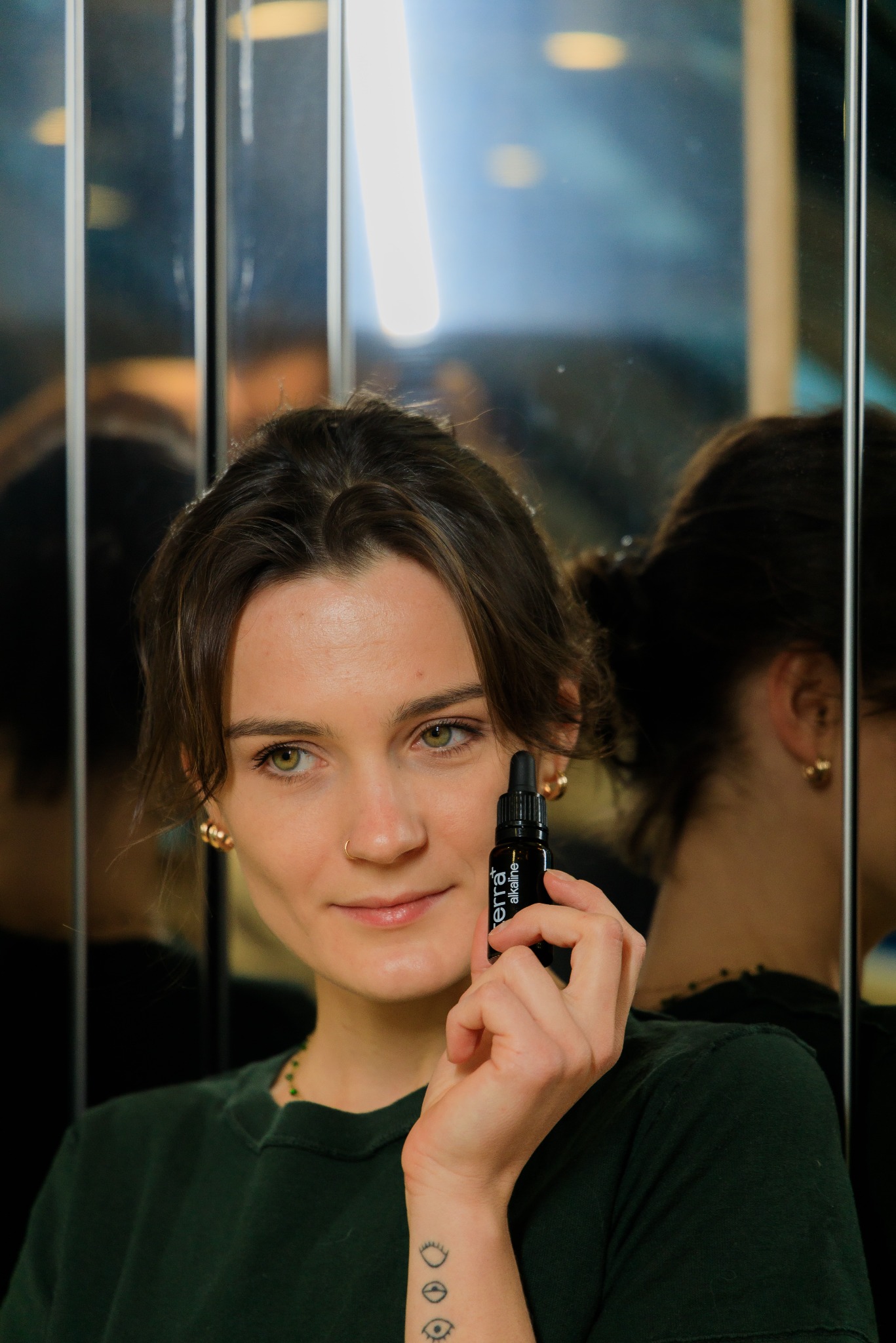 Kobieta z piegami i tatuażem oka na nadgarstku trzyma buteleczkę olejku eterycznego na tle lustrzanym.