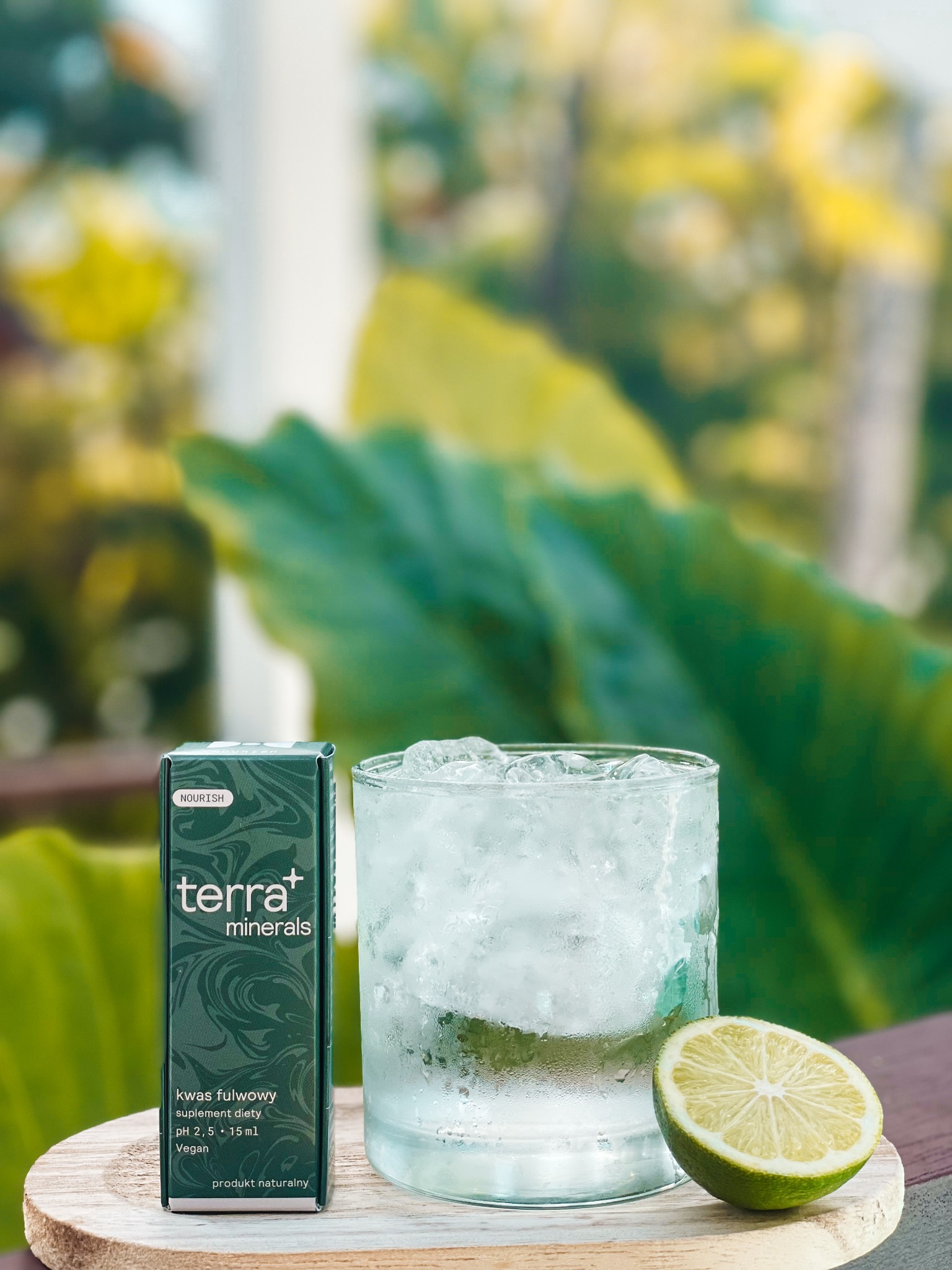 Szklanka wody z lodem i limonką obok opakowania suplementu diety kwas fulwowy na drewnianej podstawce, na tle zielonych liści.