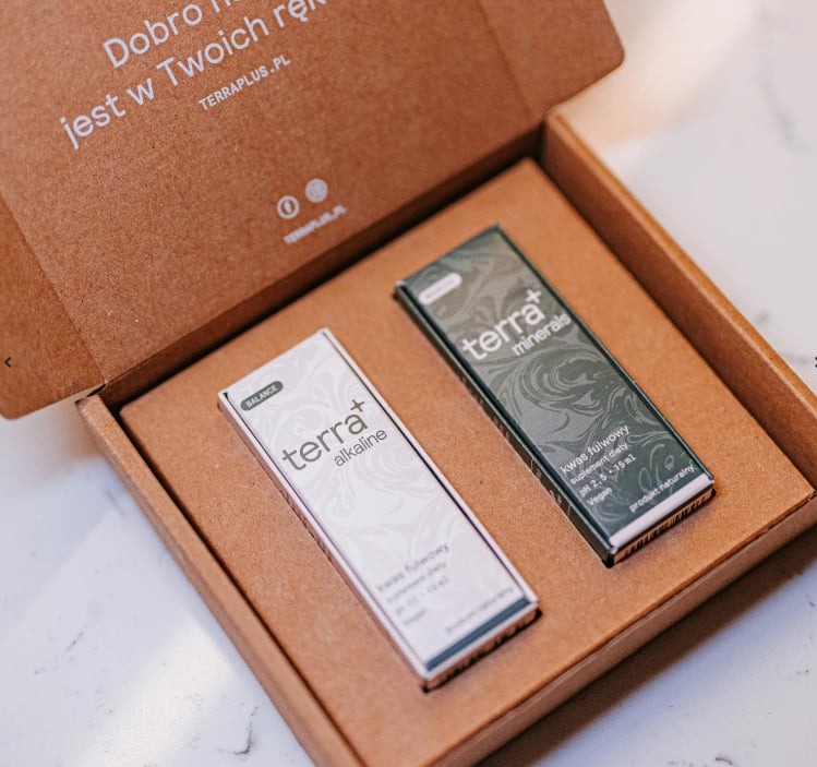 Otwarta tekturowa skrzynka prezentuje dwa opakowania suplementów diety: białe 'terra+ alkaline' i zielone 'terra+ minerals', oba z informacją o zawartości kwasu fulwowego i wegańskim składzie.