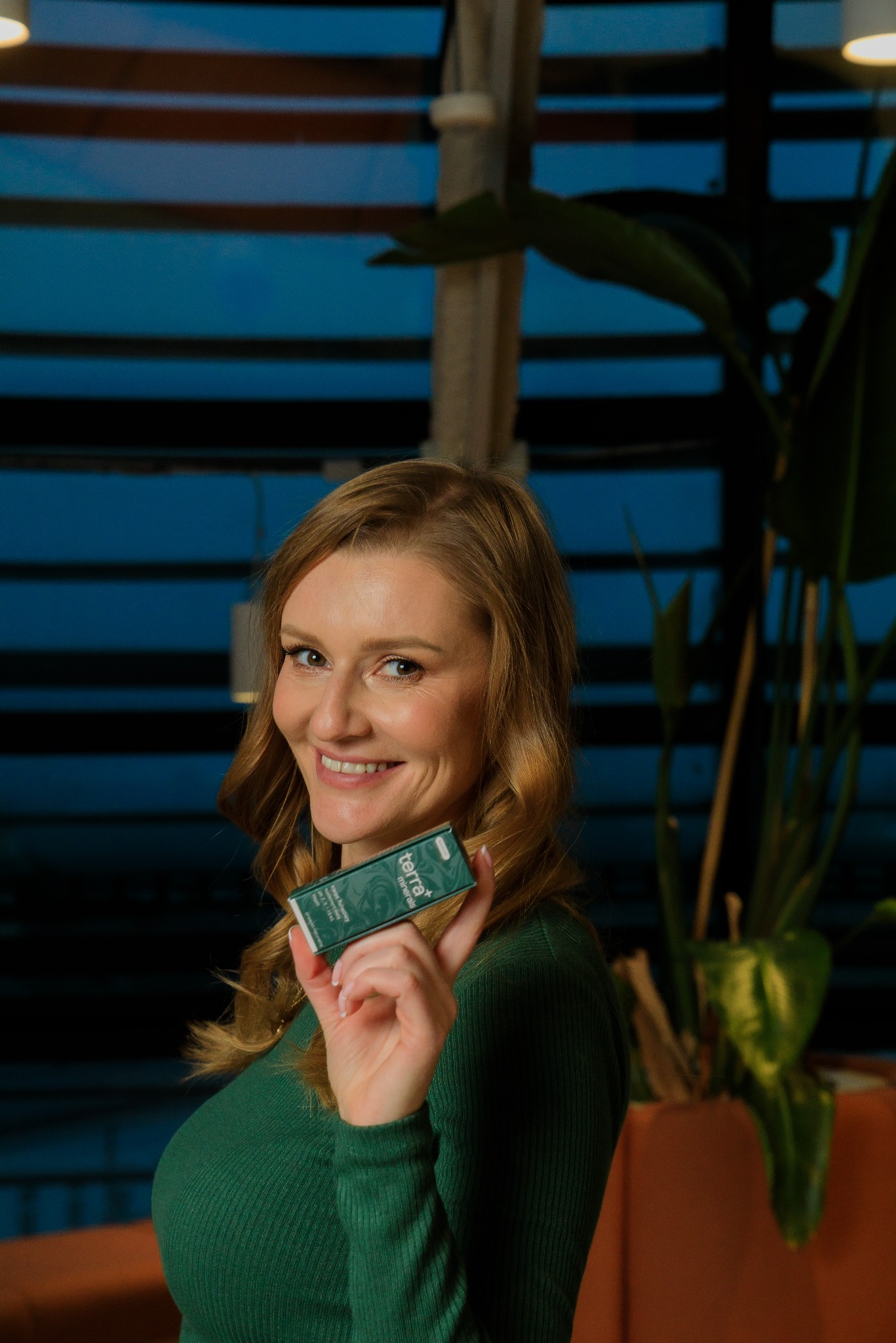 Uśmiechnięta blondynka prezentuje zielone opakowanie suplementu diety Terra+ Minerals na tle roślin i niebieskich paneli.