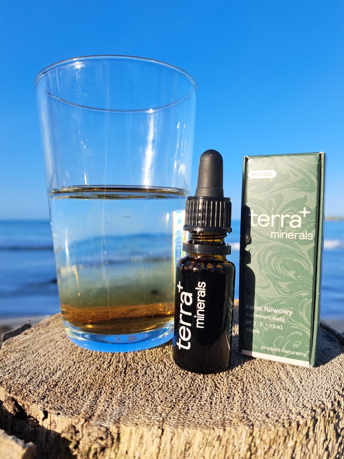 Szklanka wody z kroplami suplementu diety, obok buteleczka z pipetą i opakowanie produktu marki Terra Minerals na tle plaży i błękitnego nieba.