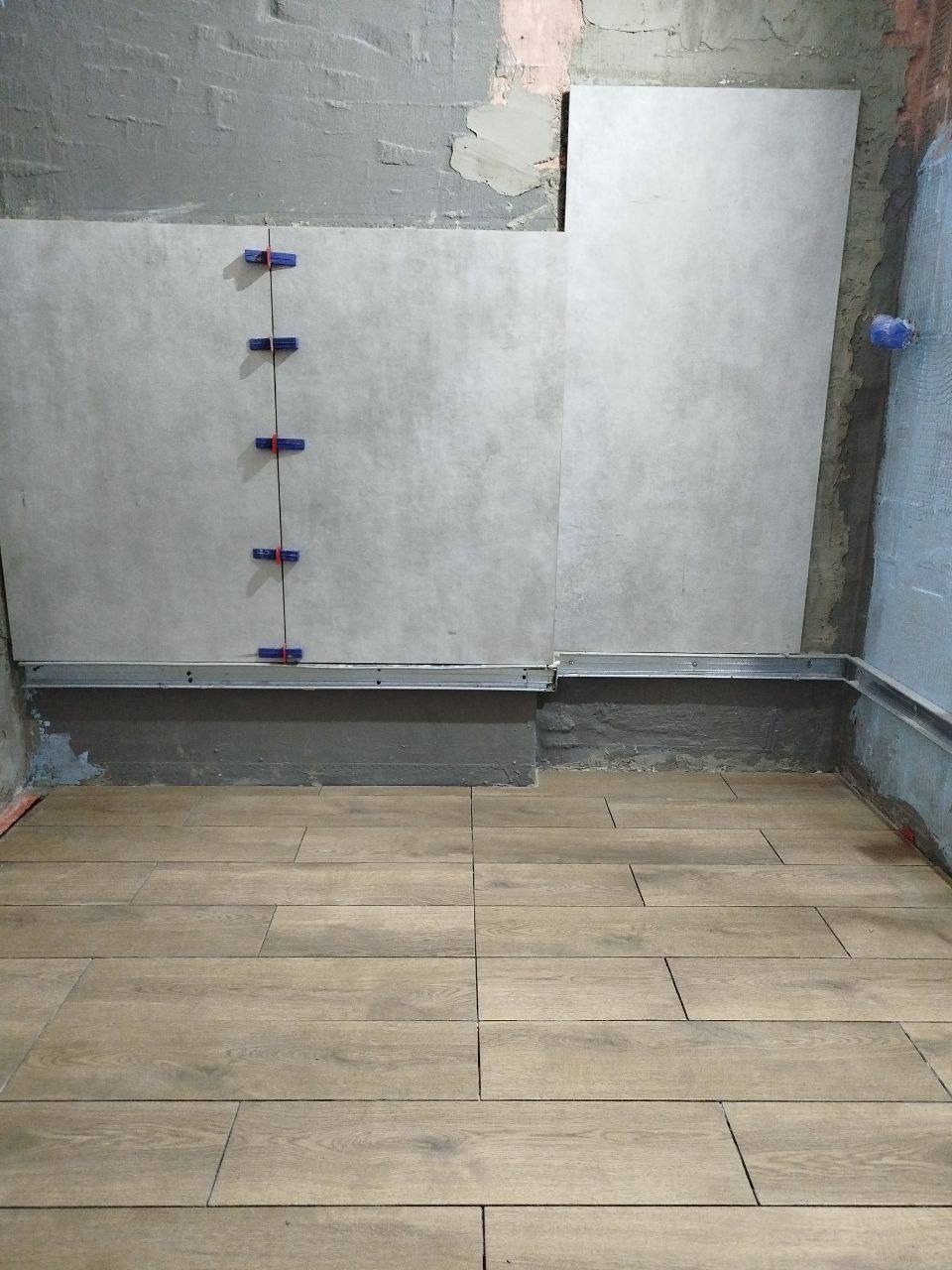 Montaż szarych płytek ściennych z użyciem klipsów poziomujących, widoczne drewnopodobne płytki podłogowe i surowa ściana z pozostałościami zaprawy.