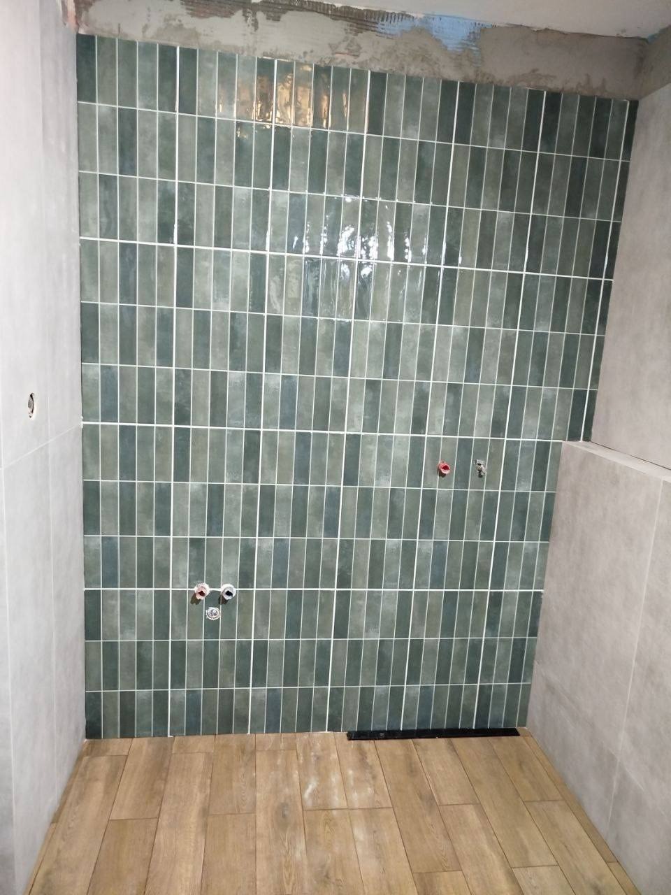 Ściana w łazience wyłożona pionowymi, zielono-szarymi płytkami z białymi fugami, drewnopodobna podłoga, widoczne przyłącza wodne i odpływ liniowy.