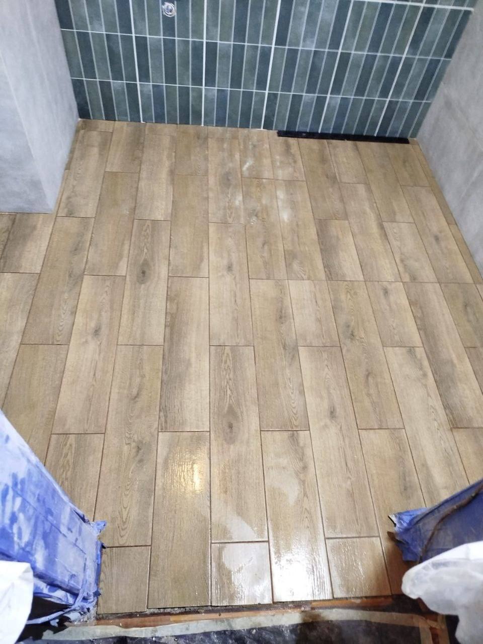 Ułożona podłoga z płytek imitujących drewno w wąskim pomieszczeniu, z szarozielonymi kafelkami na ścianie w tle.