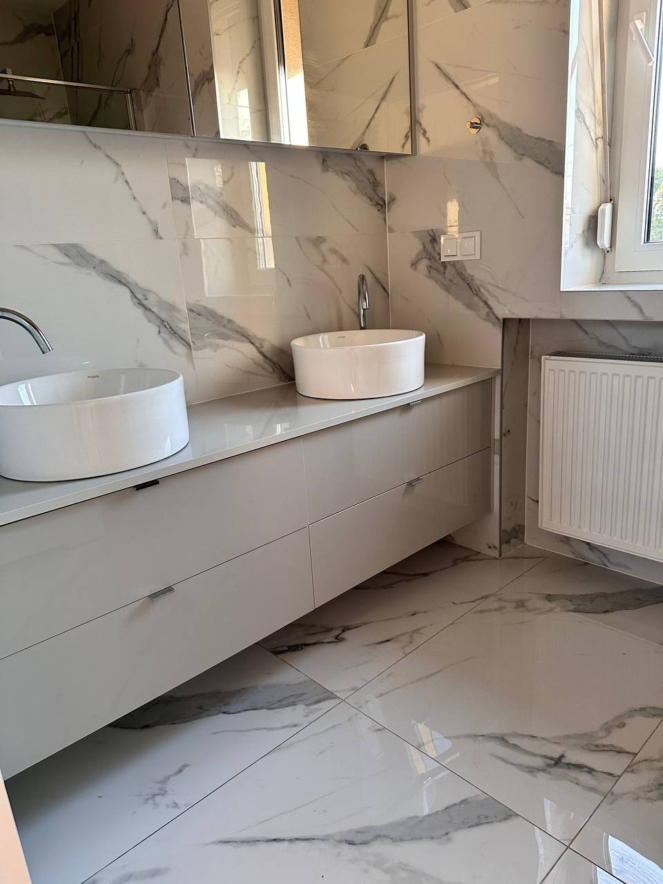 Nowoczesna łazienka z dwoma okrągłymi umywalkami nablatowymi, szafką z szufladami w jasnym odcieniu, grzejnikiem drabinkowym i marmurowymi płytkami na ścianach i podłodze.