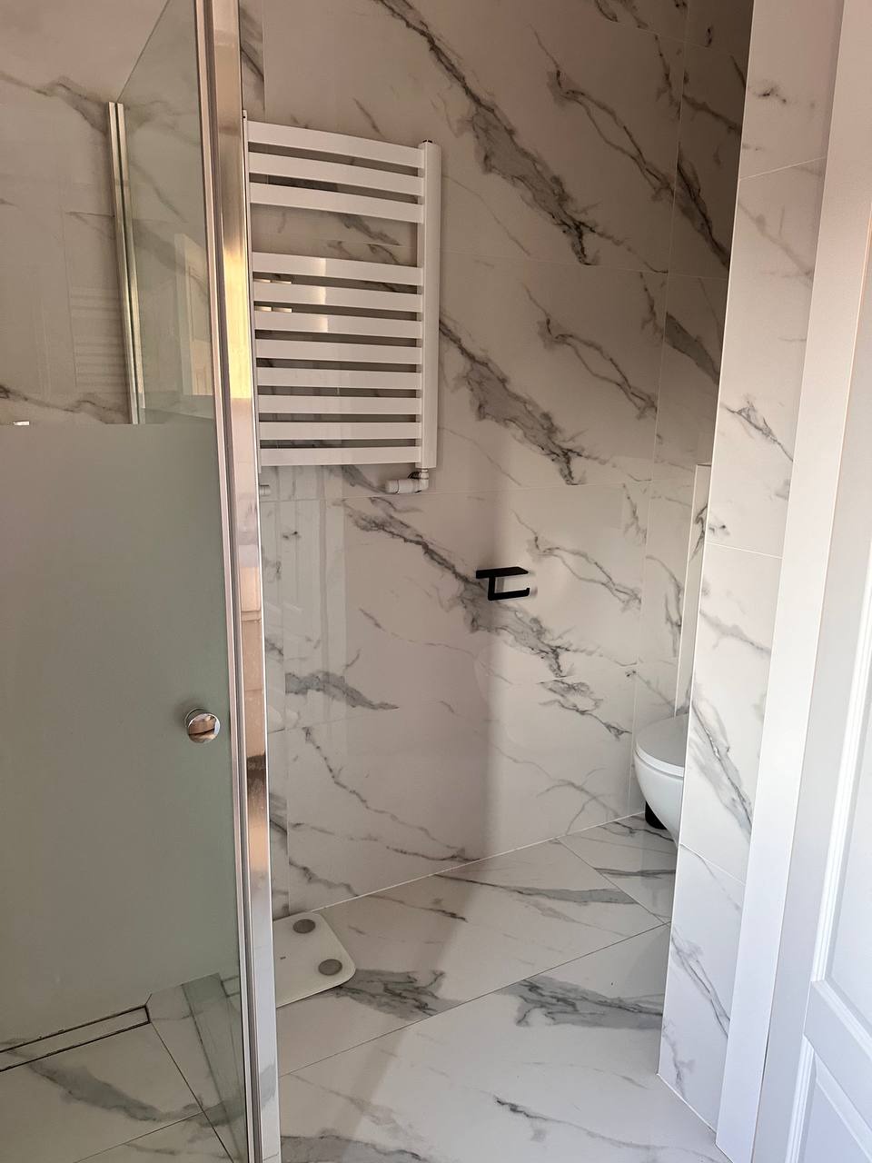 Wykończona łazienka z kabiną prysznicową, grzejnikiem drabinkowym i płytkami imitującymi marmur na ścianach i podłodze. W tle widoczna toaleta.