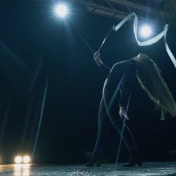 Dominik Ksel - Kobieta w czarnym stroju gimnastycznym wykonuje układ z białą wstążką na scenie w blasku reflektorów.