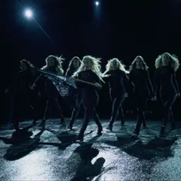 Zespół taneczny w czarnych strojach występuje na scenie oświetlonej reflektorem, jedna z tancerek trzyma flagę z napisem.