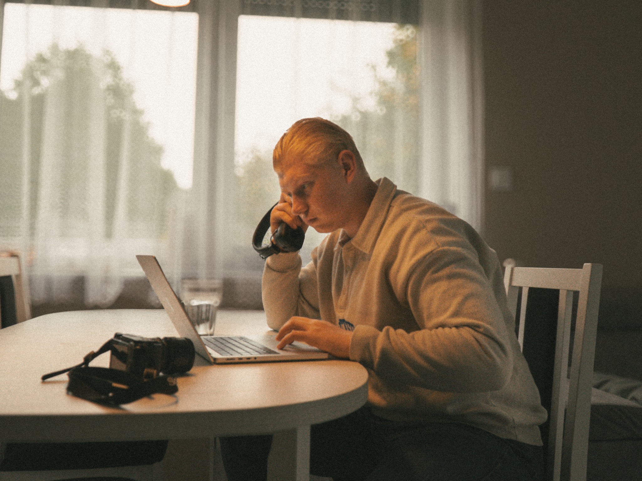 Młody mężczyzna z blond włosami, w beżowej bluzie, siedzi przy okrągłym stole i pracuje na laptopie. Na stole leży aparat fotograficzny, słuchawki na uchu i szklanka wody.